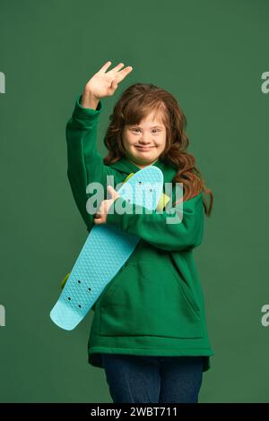 Teenager mit Down-Syndrom mit gemütlichem Hoodie, mit Skateboard vor grünem Studiohintergrund Stockfoto