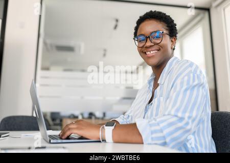 Seitenansicht einer afroamerikanischen Geschäftsfrau, die am Schreibtisch sitzt, eine lächelnde charismatische Angestellte, eine freischaffende Frau mit einem lässigen blauen Hemd und einer Brille arbeitet Stockfoto