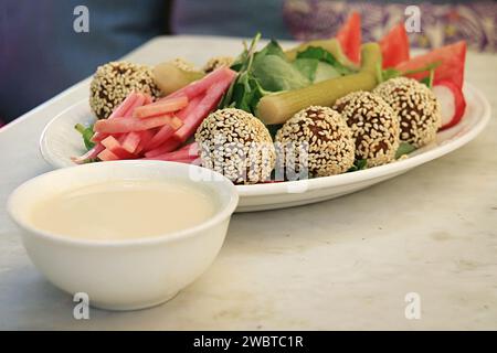 Falafelbällchen mit Sesam auf einer Platte zusammen mit dem traditionellen begleitenden Gemüse und Tahini-Sauce. Stockfoto