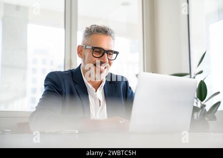 Lächelnder reifer Geschäftsmann, der formelle Anzug und Brille trägt, Laptop für die Büroarbeit benutzt, männlicher Angestellter auf den Bildschirm schaut, Nachrichten liest, Post liest Stockfoto