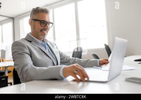 Selbstbewusster, reifer Geschäftsmann, der ein Notebook in einem Loft-Büro verwendet. Freundlicher, männlicher Mitarbeiter aus den 50er Jahren, der am Arbeitsplatz sitzt, auf den Bildschirm blickt, recherchiert, analysiert, seitlich betrachtet Stockfoto