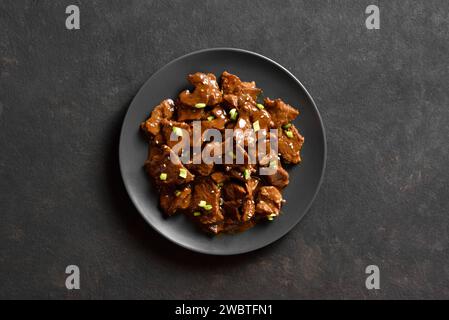 Asiatisches Rindfleisch mit Sojasauce, grünen Zwiebeln und Sesamsamen auf Teller auf dunklem Steinhintergrund. Gebratenes Rindfleisch. Draufsicht, flach Stockfoto