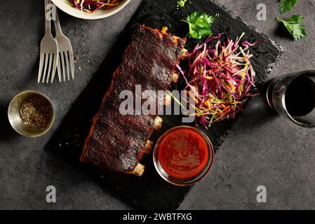 Gegrillte Schweinerippchen, serviert mit Krautsalat und Sauce auf schwarzem Schieferteller auf dunklem Hintergrund. Nahaufnahme von bbq Schweinerippchen, serviert mit lebhaften coles Stockfoto