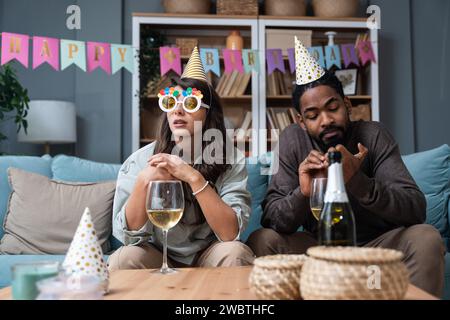 Zwei junge Leute sitzen auf Sofa mit Geburtstagskappen, traurig, weil niemand kam, um Geburtstag und Party in ihrer Wohnung zu feiern. Enttäuschter Mann und Stockfoto