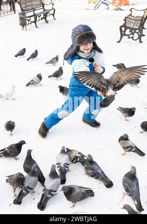 Junge mit warmem Hut und warmen Overalls verbringt an einem Wintertag in der Stadt gern Zeit umgeben von Tauben. Viele wilde Tauben fliegen in die Hände der C Stockfoto