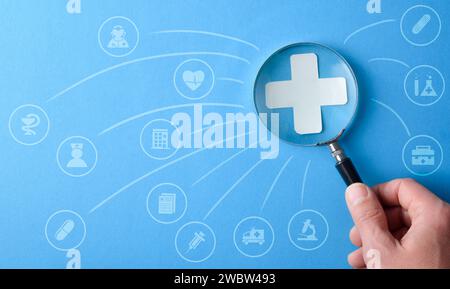 Medizin- und Versicherungsdienstkonzept mit Hand mit einer Lupe zunehmender Ausschnitt des medizinischen Kreuzes mit verschiedenen Dienstleistungssymbolen auf blauem Hintergrund Stockfoto