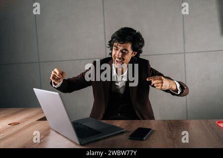 Glücklicher, aufgeregter Geschäftsmann im Anzug, der geschäftliche Erfolge feiert und auf den Laptop-Bildschirm blickt. Fröhlich lächelnder Mitarbeiter, der sich motiviert fühlt Stockfoto