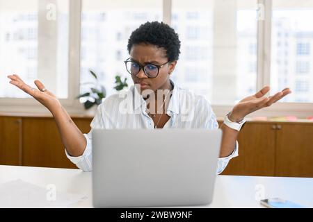 Verwirrte afroamerikanische Büroangestellte, die am Arbeitsplatz sitzt und mit den Schultern zuckt, Probleme mit dem Projekt hat, Computerfehler hat, Geschäftsfrau in formeller Kleidung, die auf den Computermonitor schaut Stockfoto
