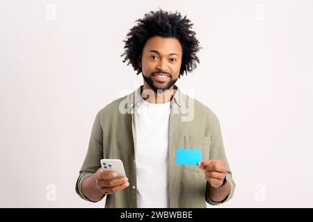 Fröhlicher Kerl, der Smartphone und Kreditkarte benutzt, um online einzukaufen. Glücklicher Mann bestellt online Essen, männlich bezahlt für langersehnten Kauf auf dem Handy, isoliert auf weiß Stockfoto