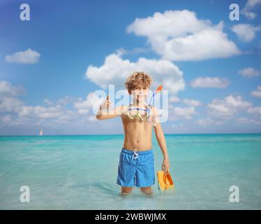 Junge, der im Meer steht und eine Tauchermaske hält und Daumen nach oben bewegt Stockfoto