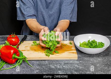 Grüne Salatblätter in den Händen in der Nähe der weißen Schüssel. Blaues T-Shirt, schwarzer Hintergrund. Helles Gemüse Stockfoto