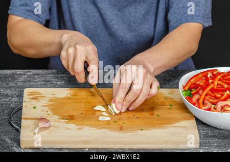 Hände einer Frau, die frischen Knoblauch auf Holzbrett hackt. Schälsalat auf grauem Tisch, schwarzer Hintergrund Stockfoto