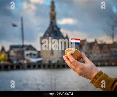 Oliebol mit niederländischer Flagge in der Hand, traditionelle niederländische Leckereien, die normalerweise im Winter auf dem Hintergrund des Hafens der Stadt Hoorn gegessen werden Stockfoto