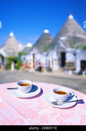 Nahaufnahme von zwei Tassen Espresso auf einem Tisch mit rosa Tischdecke, mit leerer Straße und Souvenirläden im Hintergrund in Alberobello in PR Stockfoto