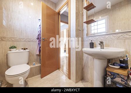 Ein kleines Badezimmer mit Fliesenwänden in Marmor-Optik, einem Spiegel mit abgeschrägten Kanten, der in die Wand integriert ist, und einem Waschbecken mit passendem Porzellansockel Stockfoto