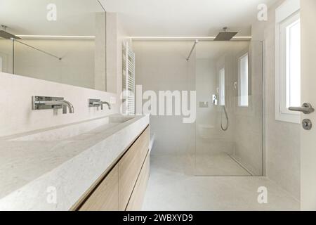 Modernes Design-Badezimmer mit in die Wand integriertem Spiegel, cremefarbenen Fliesenwänden und -Böden, Eichenmöbeln mit mehreren Schubladen und einer Sitzeinrichtung Stockfoto
