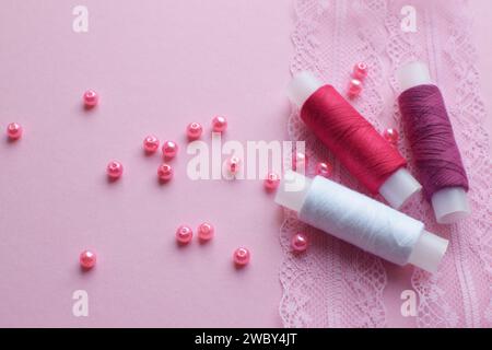 Rote und weiße Baumwollfäden in Spulen und rosa Perlen liegen auf rosa Spitze. Blick von oben Stockfoto