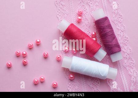 Rote und weiße Baumwollfäden in Spulen und rosa Perlen liegen auf rosa Spitze. Blick von oben Stockfoto