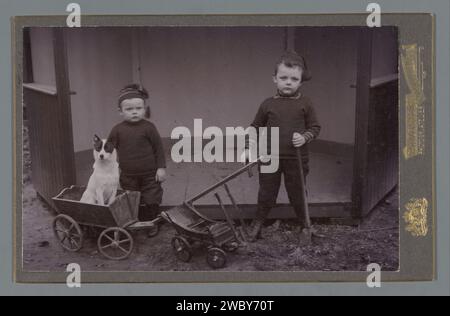 Porträt zweier Jungen mit Strickmützen auf Spielzeugwagen und einem Hund, C. Sanders SZN, ca. 1900 - 1930 Foto. Schrankfoto mit Gartenschuppen im Hintergrund. Unterstützung für Fotos. Kind aus Karton (zu Hause). Hund. Spielzeuge. Zusammenfügen Stockfoto
