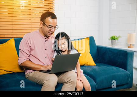 Ein lächelnder asiatischer Vater arbeitet von zu Hause aus auf seinem Laptop, während seine Tochter auf einem Computer für e lernt Stockfoto
