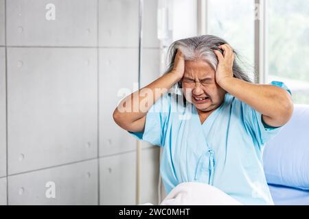 Eine ältere asiatische Frau mit starken Kopfschmerzen, die in einem Krankenhausbett sitzt Stockfoto