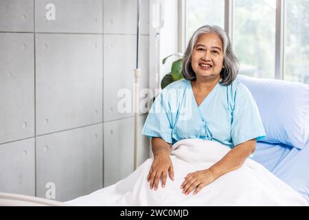 Eine ältere, lächelnde asiatische Frau, die auf einem ordentlich gemachten Krankenhausbett in einem Krankenhaus sitzt Stockfoto