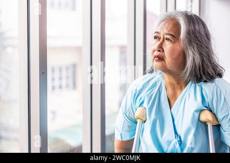 Eine asiatische, nachdenkliche Frau, die sich in einem Krankenhaus auf Krücken stützt und aus einem Fenster blickt Stockfoto