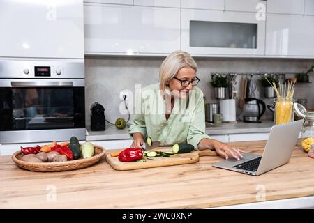 Ältere Frau eine Witwe kocht vegetarische gesunde Lebensmittel aus Gemüse zu Hause in der Küche, mit Laptop-Computer für Online-Rezepte, Vorbereitung Stockfoto