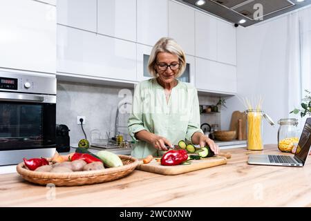 Ältere Frau eine Witwe kocht vegetarische gesunde Lebensmittel aus Gemüse zu Hause in der Küche, mit Laptop-Computer für Online-Rezepte, Vorbereitung Stockfoto