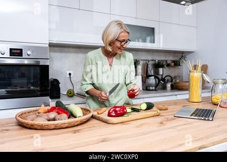 Ältere Frau eine Witwe kocht vegetarische gesunde Lebensmittel aus Gemüse zu Hause in der Küche, mit Laptop-Computer für Online-Rezepte, Vorbereitung Stockfoto