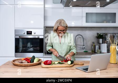 Ältere Frau eine Witwe kocht vegetarische gesunde Lebensmittel aus Gemüse zu Hause in der Küche, mit Laptop-Computer für Online-Rezepte, Vorbereitung Stockfoto