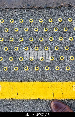 Taktiler Warnstreifen, gelbe Linie mit schwarzen und gelben Nieten am Bahnsteigrand, Vereinigtes Königreich, mit einem Teil des Pendlerschuhs sichtbar Stockfoto
