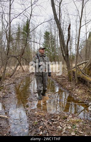 Ein Jäger bahnt sich seinen Weg durch eine Pfütze im Frühlingswald. Er hat eine Schrotflinte über die Schulter geschleudert. Die Abenddämmerung wird tiefer. Stockfoto