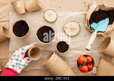 Pflanzen von Tomatensamen für Setzlinge, Hände mit einem Topf, umweltfreundliche Töpfe und Samen, Mahl- und Gartenwerkzeuge auf Bastelpapier, Vorbereitung mit Frühling Stockfoto