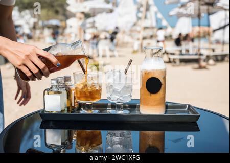 Schwarzer Kaffee von Hand aus der Flasche in Glas mit Eiswürfel und Eislatte, Sirup auf Tablett an sonnigem Tag Stockfoto