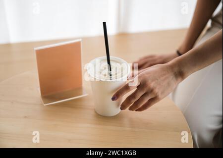 Hand einer Frau, die Einweg-Kaffeetasse auf Holztisch mit leerem Etikett im Café hält Stockfoto