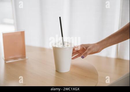 Hand einer Frau, die Einweg-Kaffeetasse auf Holztisch mit leerem Etikett im Café hält Stockfoto