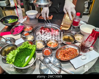 Das Personal grillt im koreanischen Restaurant geschnittenes mariniertes Schweinefleisch auf einem Holzkohleofen mit koreanischen Beilagen und Gurken Stockfoto