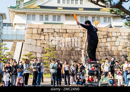 Ein Jongleur balanciert auf kleinen Tischen und Trommeln und unterhält eine kleine Menschenmenge, die an einem Frühlingstag im Osaka Castle Park im hellen Sonnenschein beobachtet. Stockfoto