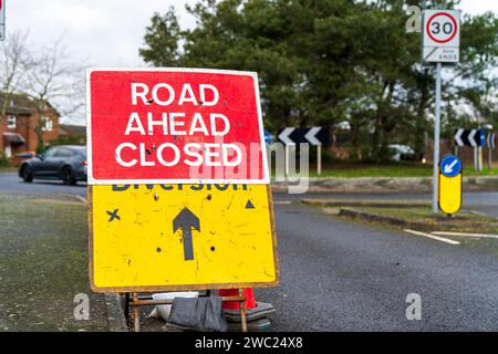 Britisches rotes Schild „Road Ahead Closed“ mit einem weiteren gelben Schild, „Diversion“ mit schwarzem Kreuz und Dreieck, das angibt, welche Seite geschlossen ist. Stockfoto