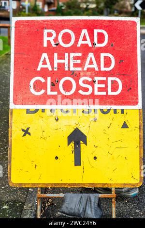 Britisches rotes Schild „Road Ahead Closed“ mit einem weiteren gelben Schild, „Diversion“ mit schwarzem Kreuz und Dreieck, das angibt, welche Seite geschlossen ist. Stockfoto