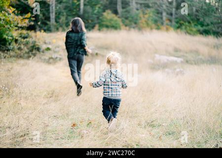 Das kleine Mädchen rennt ihrer Mutter auf dem Herbstrasen im Wald nach. Rückansicht Stockfoto