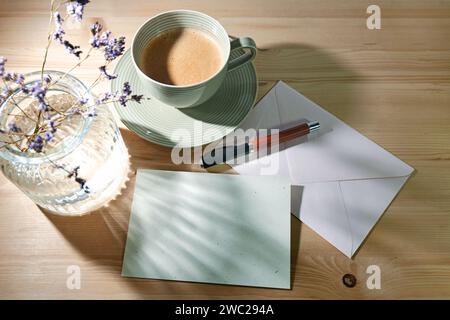 Leere Grußkarte und Umschlag aus Naturpapier, Stift, Kaffeetasse und Glasvase mit sanften Sonnenlichtreflexionen auf einem Holztisch, pastellfarben Stockfoto