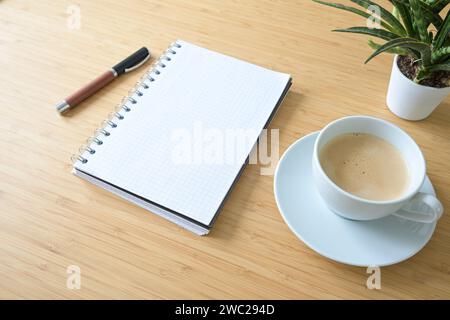 Leeres Spiralnotizblock, Kaffeetasse, Stift und eine Pflanze auf einem Schreibtisch aus Bambusholz, Business- oder Home Office-Konzept, Mockup, Kopierraum, ausgewählter Fokus, Stockfoto