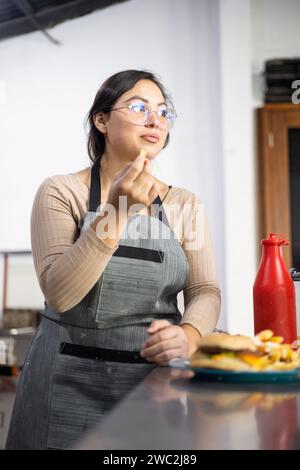 Eine junge Frau isst knusprige pommes. Teller mit Cheeseburger, Lebensmittelzubereitung in der Küche, Lebensstil am Arbeitsplatz Stockfoto