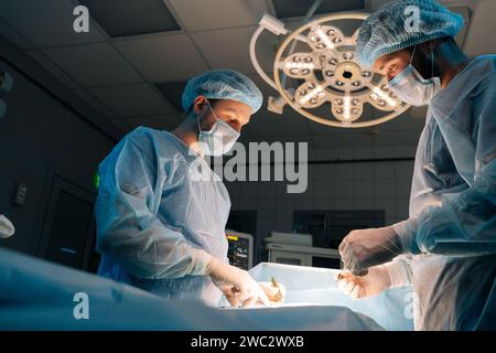 Niederwinkelansicht zweier erfahrener Chirurgen mit sterilen Handschuhen, chirurgischen Uniformen und Masken bei der chirurgischen Operation im dunklen Operationssaal Stockfoto