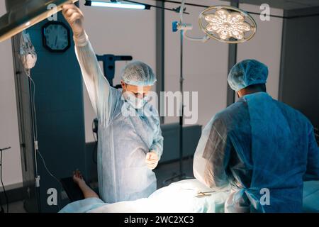 Seitliche Sicht auf die Operation durch professionelle Chirurgen. Männlicher Arzt, der das Licht einstellt und Operation mit medizinischem Kleid durchführt. Stockfoto