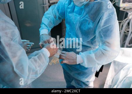 Nahaufnahme eines unerkennbaren Ärzteteams, der auf die Operation vorbereitet ist. Nahaufnahme einer Krankenschwesterassistentin, die dem Chirurgen beim Anziehen weißer Handschuhe hilft. Stockfoto