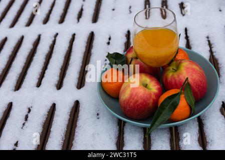 Glas frisch gepressten Bio-Fruchtsaft auf schneebedecktem Tisch im Garten und Mandarinen mit Äpfeln in der Schüssel. Gesunder Lebensstil Hintergrund. Stockfoto