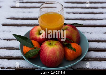 Glas frisch gepressten Bio-Fruchtsaft auf schneebedecktem Tisch im Garten und Mandarinen mit Äpfeln in der Schüssel. Gesunder Lebensstil Hintergrund. Stockfoto